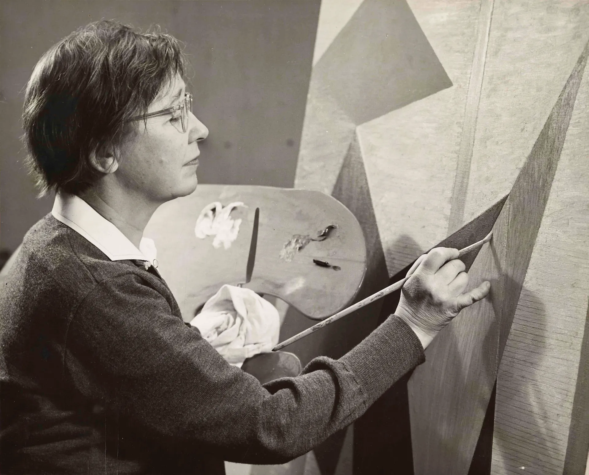 Alice Trumbull Mason painting “Mobile Tensions,” 1954 A black and white photograph of a light-skinned woman, holding a palette in her left hand and a paint brush on the right; she is in the process of creating an abstract painting.