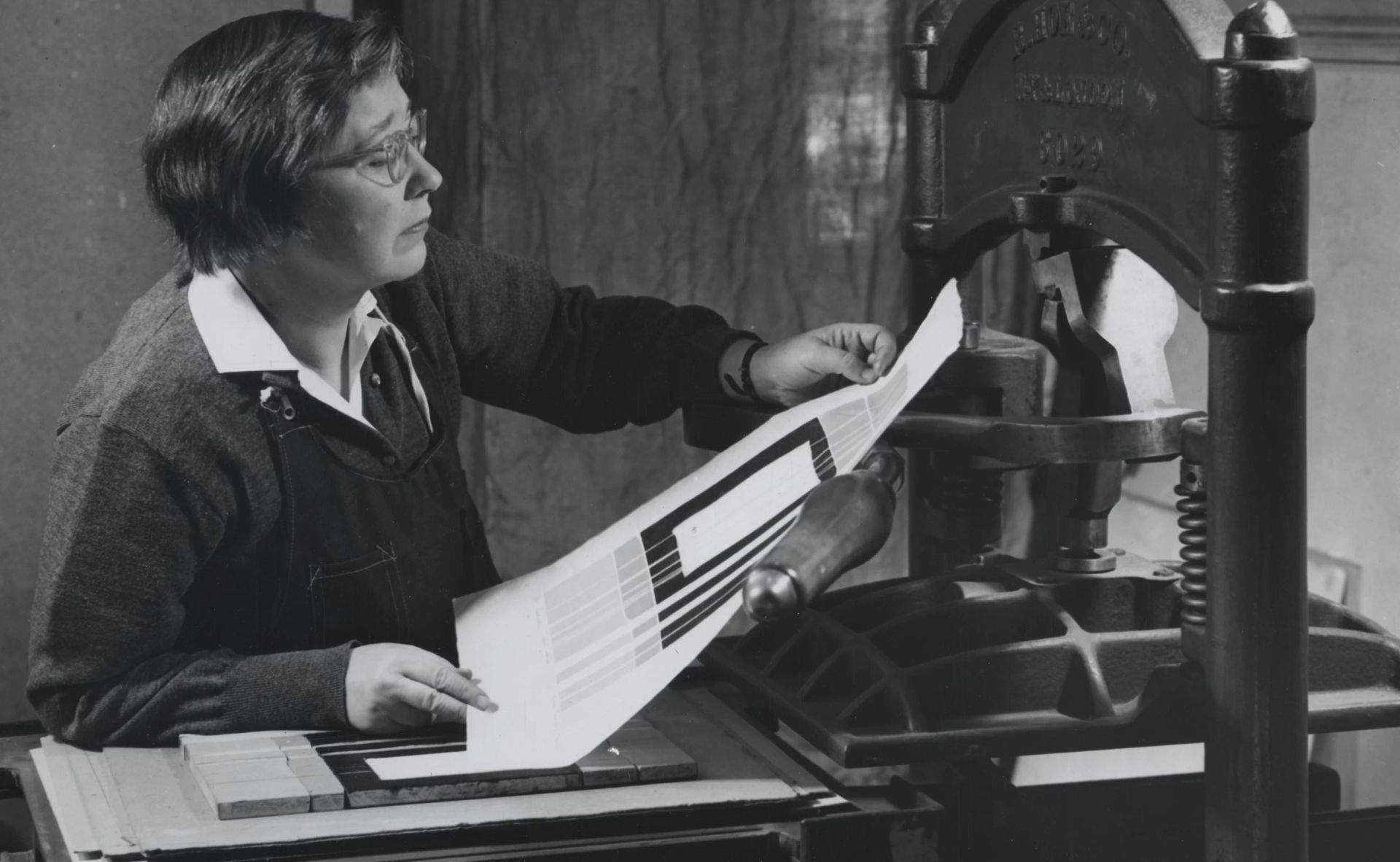 Mason in 1954 pulling her woodcut “Tidal Depths, Sekishu” off of the press. Photograph by John D. Schiff Black and white photograph from 1954 of women a woman with pale skin, examining a print just pulled off of a large printmaking press.
